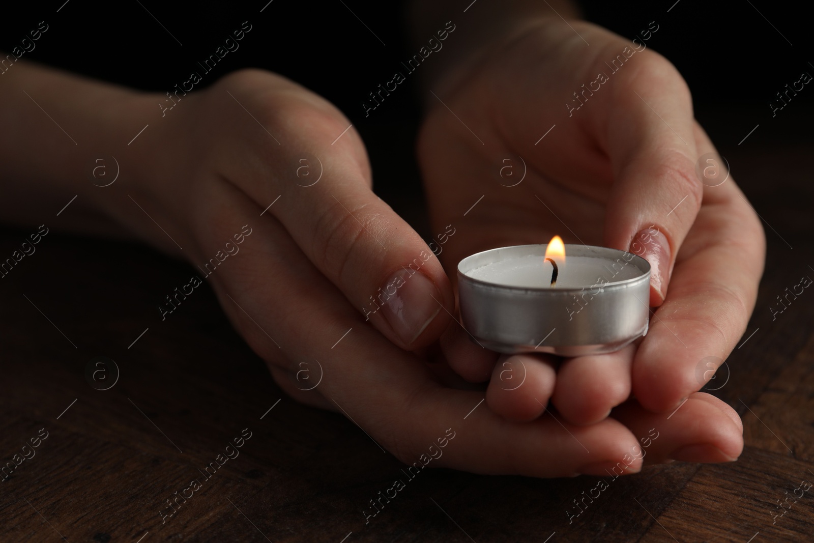 Photo of Woman holding burning tealight candle at wooden table, closeup