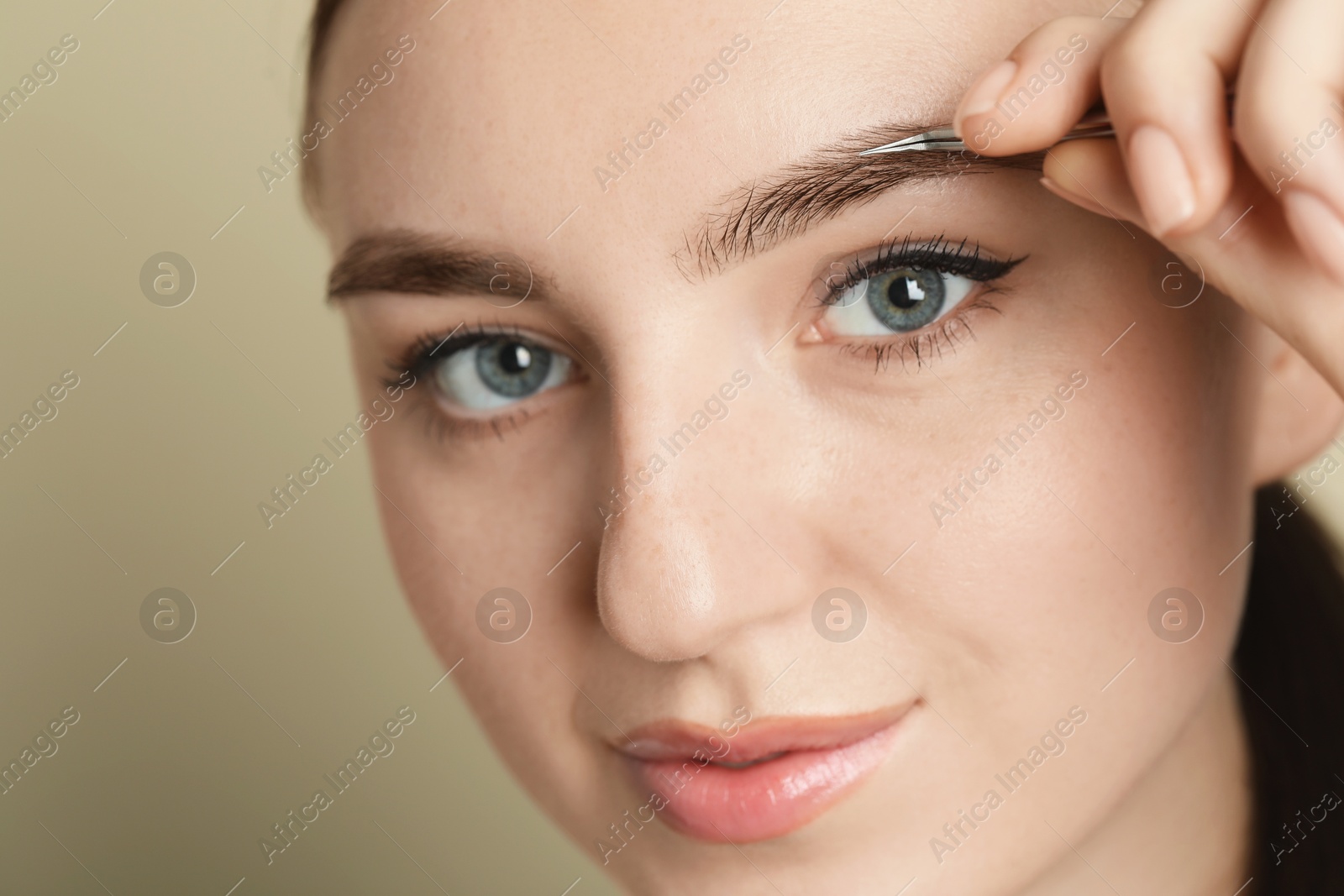 Photo of Young woman plucking eyebrow with tweezers on beige background, closeup