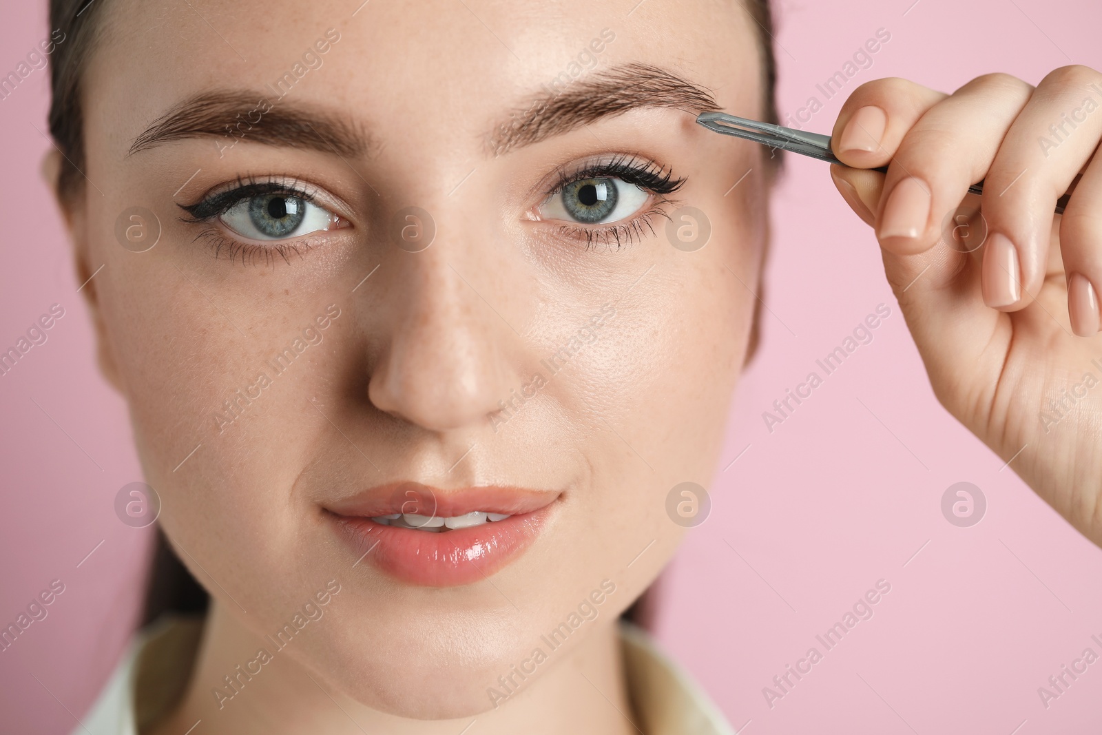 Photo of Young woman plucking eyebrow with tweezers on pink background, closeup