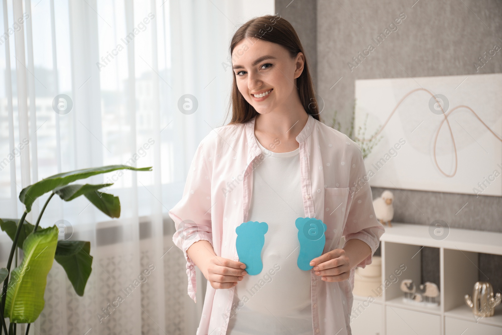 Photo of Expecting twins. Pregnant woman holding two paper cutouts of feet at home