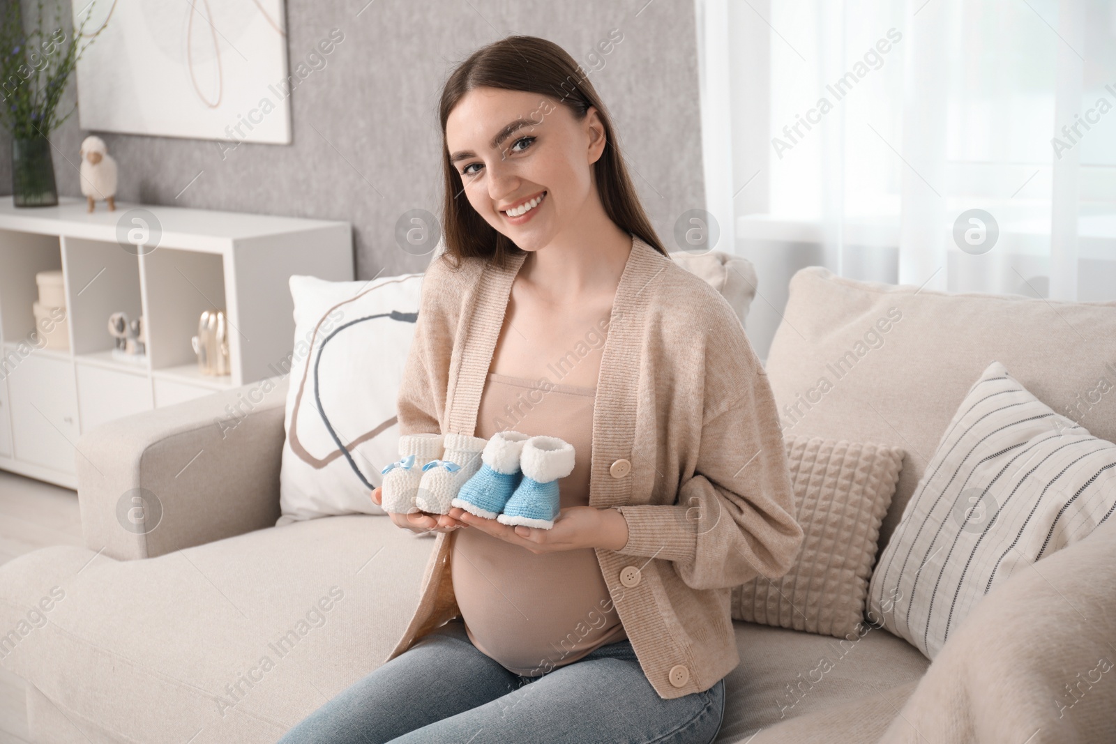 Photo of Expecting twins. Pregnant woman holding two pairs of shoes at home