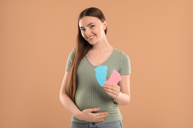 Photo of Expecting twins. Pregnant woman holding two paper cutouts of feet on light brown background