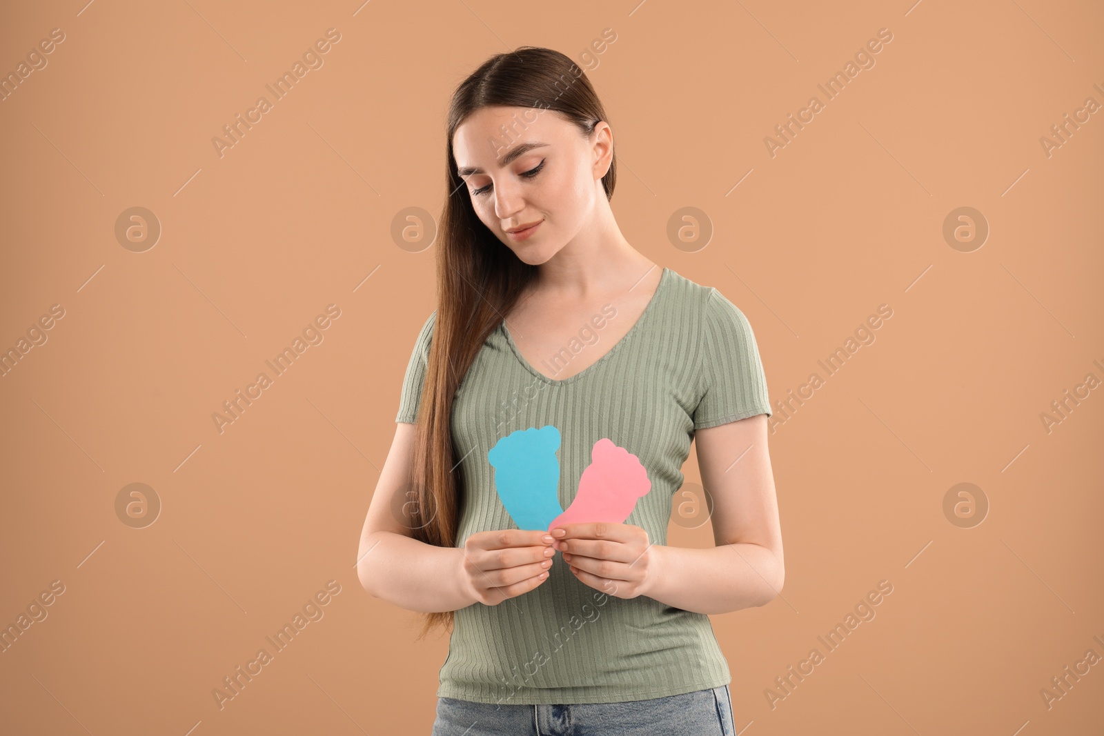 Photo of Expecting twins. Pregnant woman holding two paper cutouts of feet on light brown background