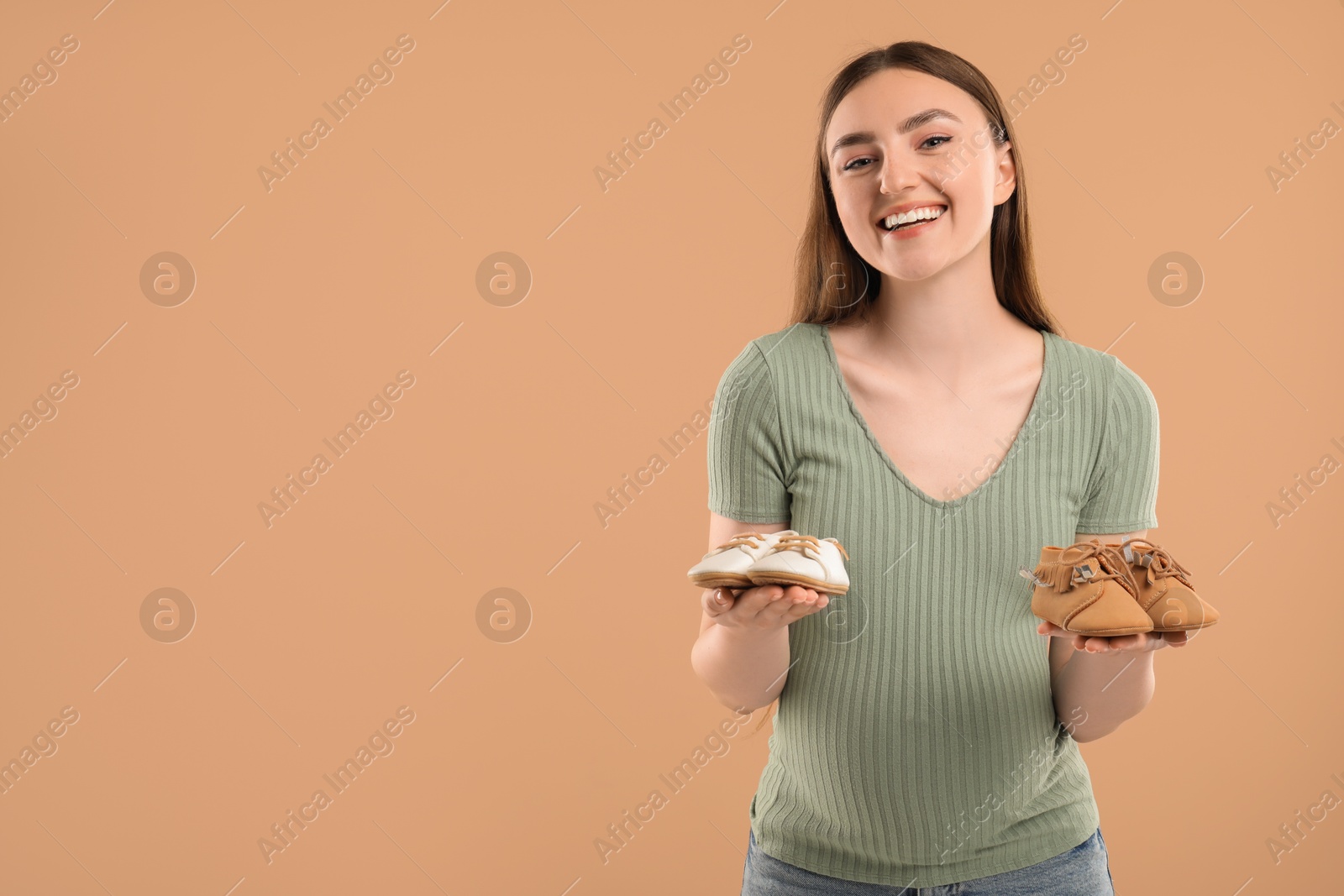 Photo of Expecting twins. Pregnant woman holding two pairs of shoes on light brown background, space for text