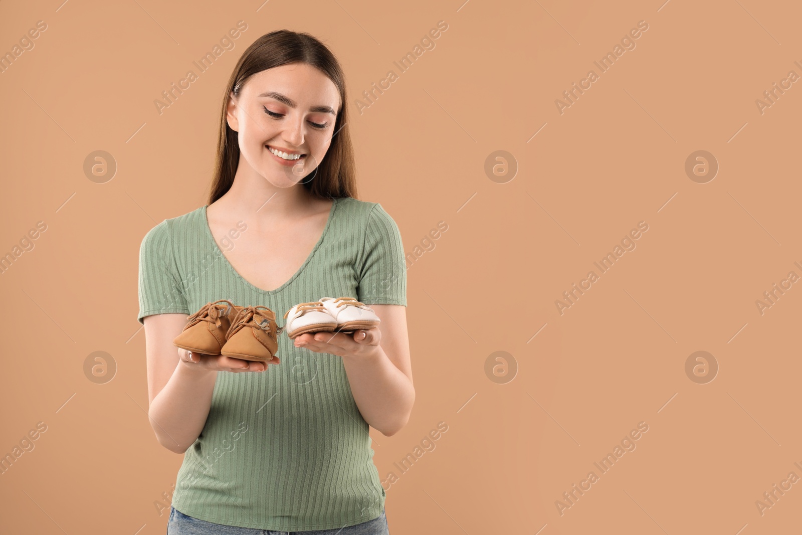 Photo of Expecting twins. Pregnant woman holding two pairs of shoes on light brown background, space for text