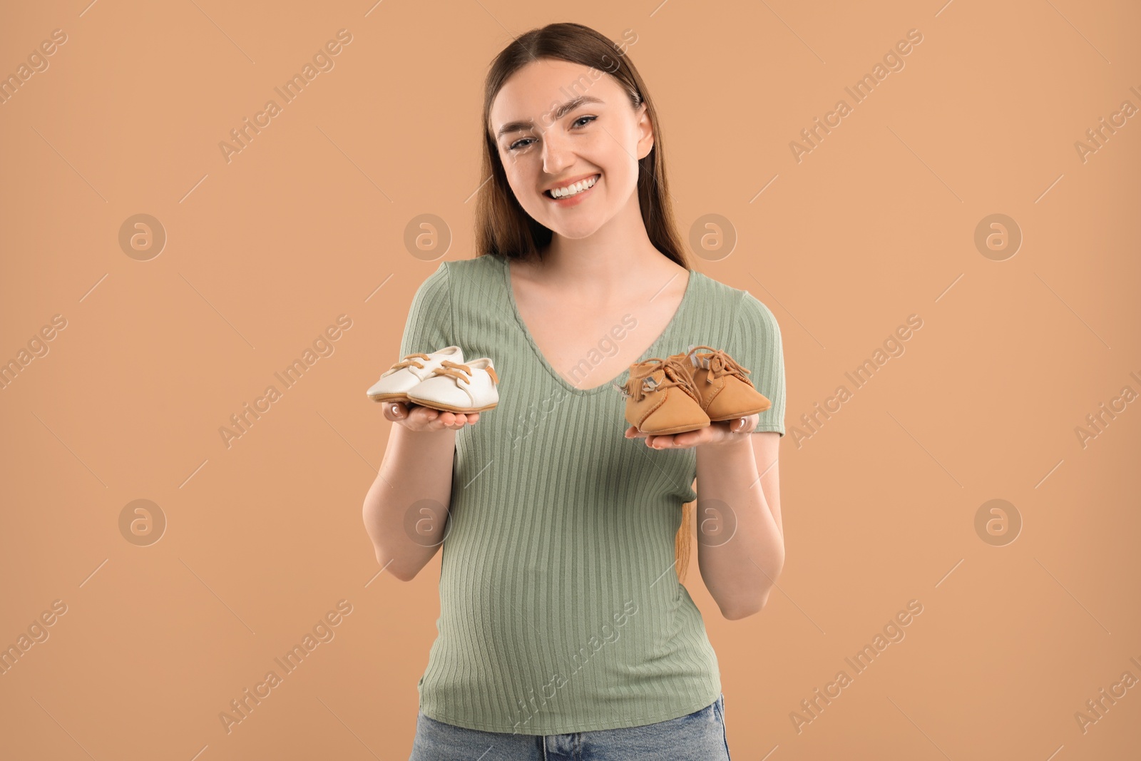 Photo of Expecting twins. Pregnant woman holding two pairs of shoes on light brown background