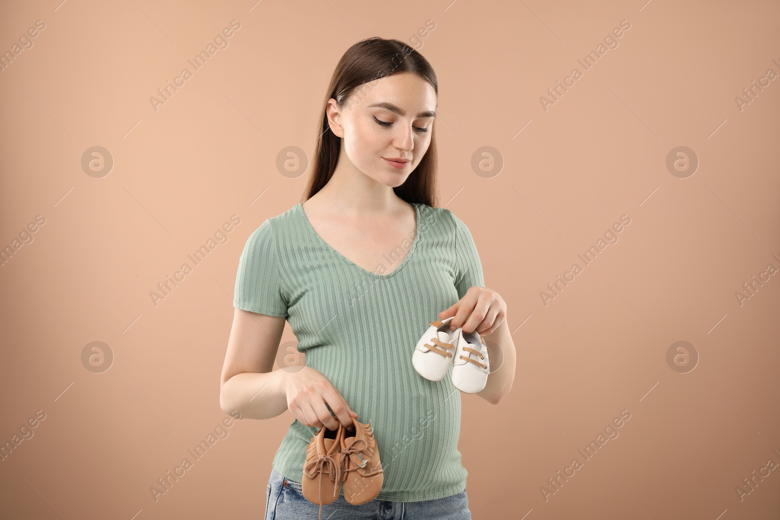 Photo of Expecting twins. Pregnant woman holding two pairs of shoes on light brown background