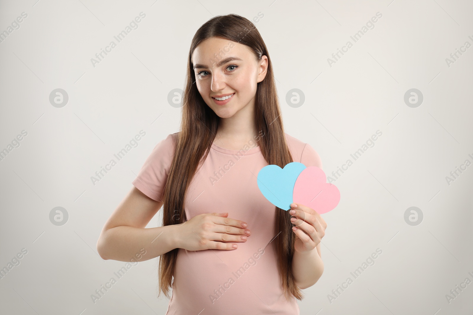 Photo of Expecting twins. Pregnant woman holding two paper cutouts of hearts on light grey background