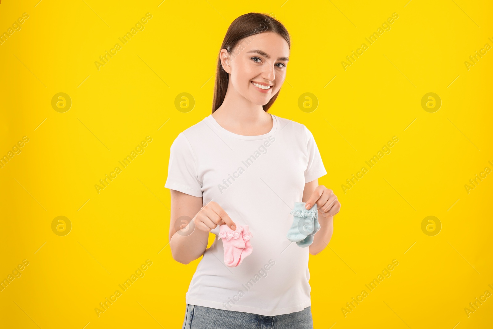 Photo of Expecting twins. Pregnant woman holding two pairs of socks on yellow background