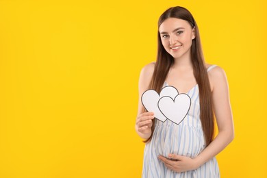 Photo of Expecting twins. Pregnant woman holding two paper cutouts of hearts on yellow background, space for text