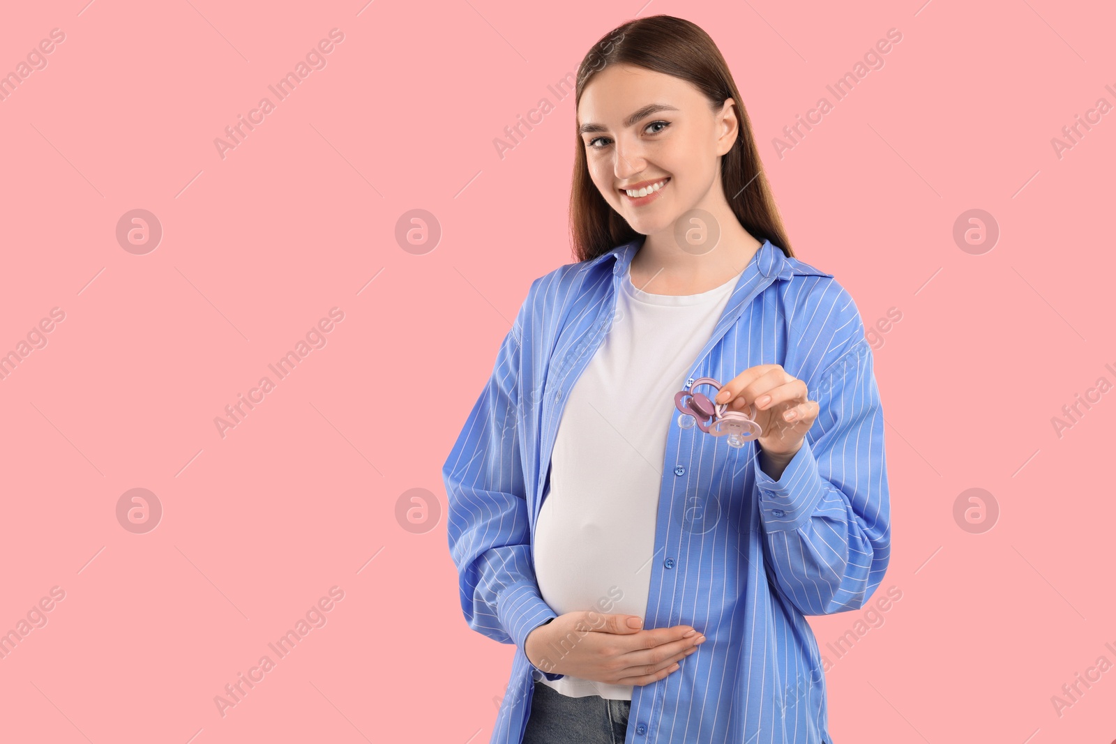 Photo of Expecting twins. Pregnant woman holding two pacifiers on pink background, space for text