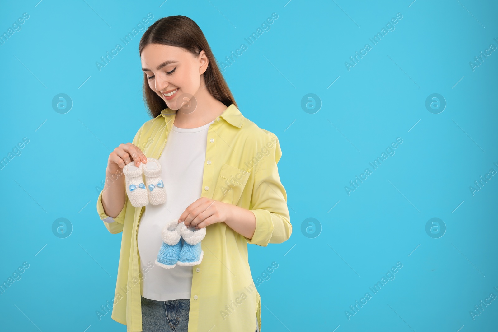 Photo of Expecting twins. Pregnant woman holding two pairs of baby shoes on light blue background, space for text