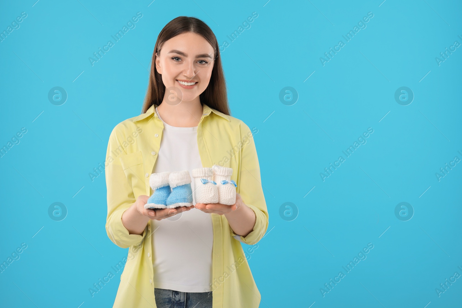 Photo of Expecting twins. Pregnant woman holding two pairs of baby shoes on light blue background, space for text