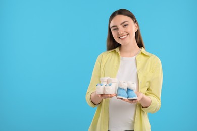 Photo of Expecting twins. Pregnant woman holding two pairs of baby shoes on light blue background, space for text