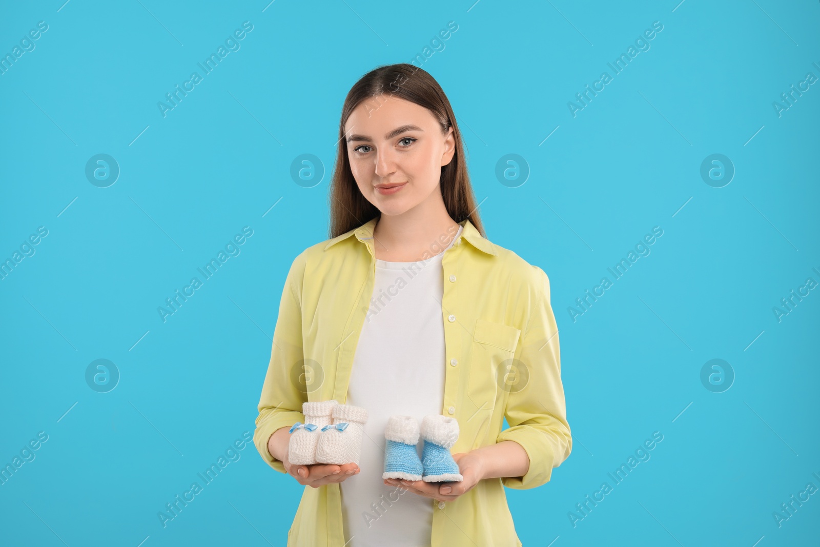 Photo of Expecting twins. Pregnant woman holding two pairs of baby shoes on light blue background