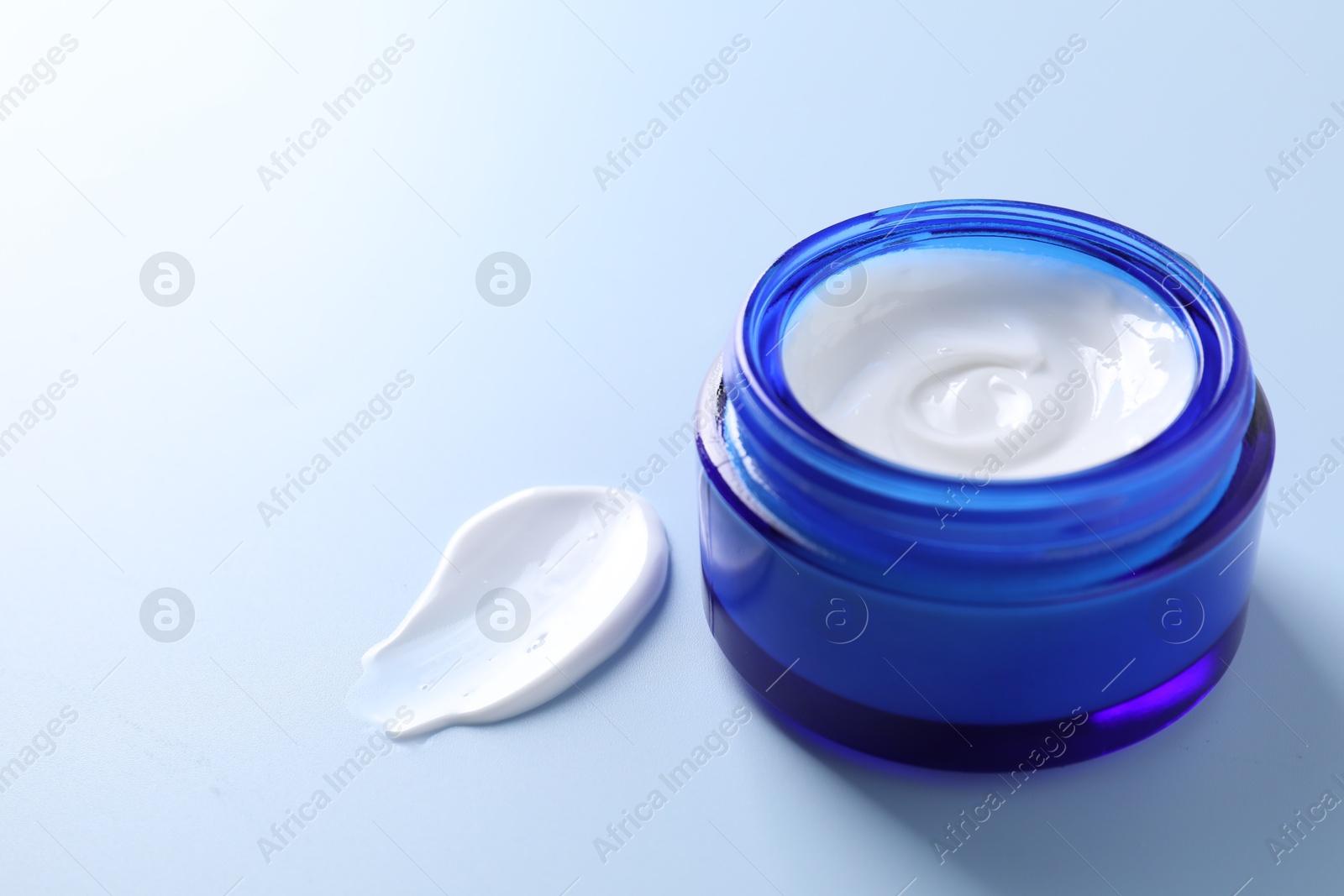 Photo of Face cream in jar and smear on light blue background, closeup. Space for text