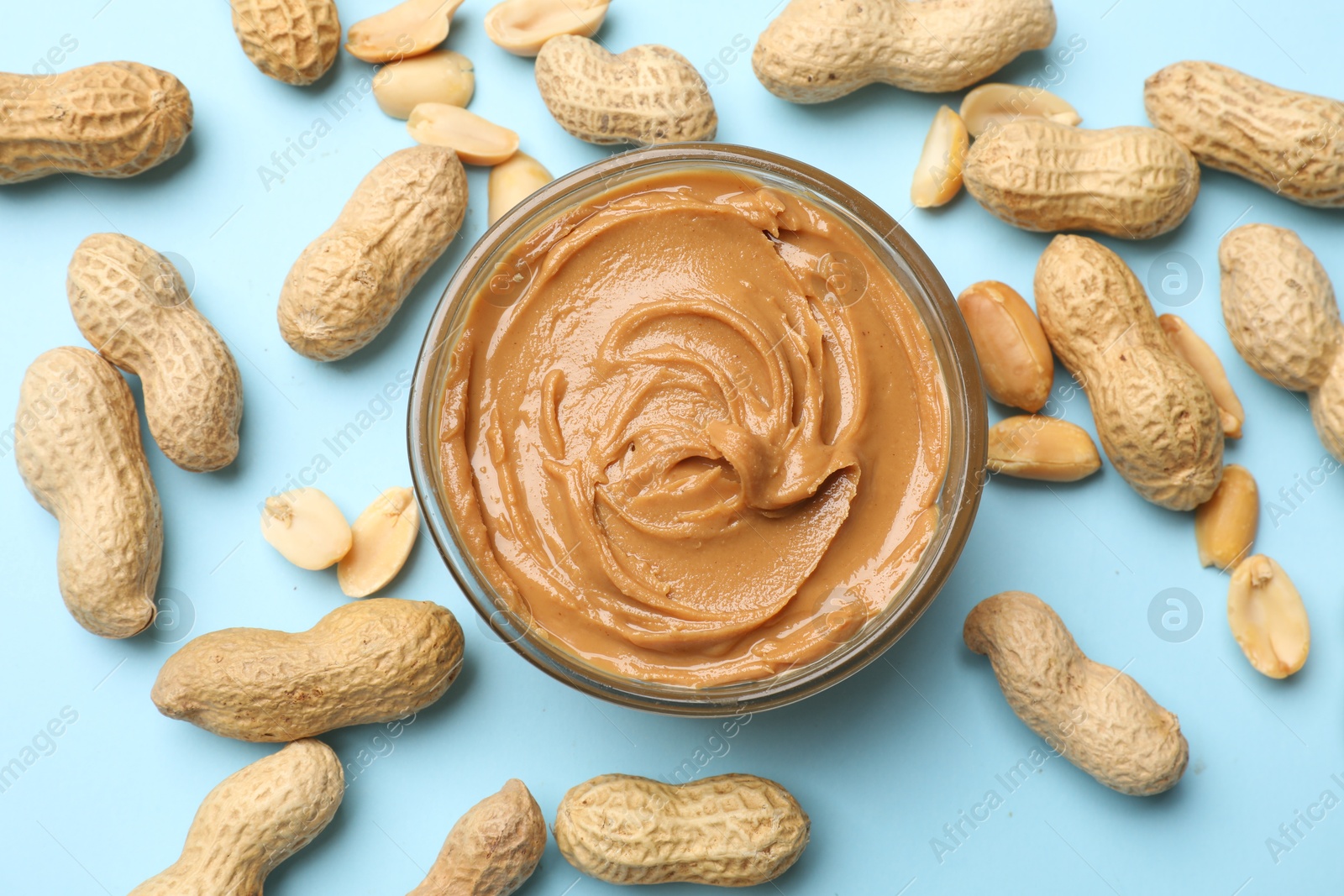 Photo of Tasty peanut butter in bowl and groundnuts on light blue background, flat lay