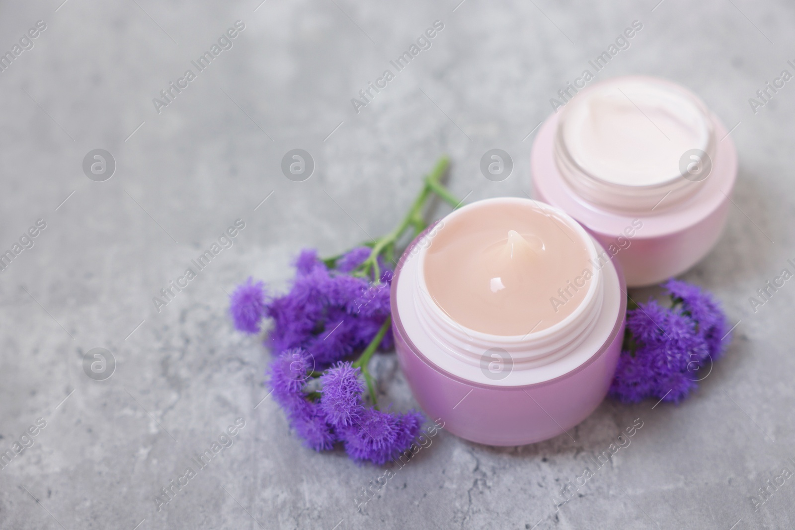 Photo of Jars of creams and ageratum flowers on gray table, closeup. Space for text