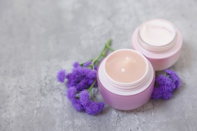 Photo of Jars of creams and ageratum flowers on gray table, closeup. Space for text