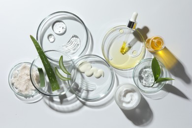 Photo of Petri dishes with different cosmetic products and aloe vera leaves on white background, flat lay