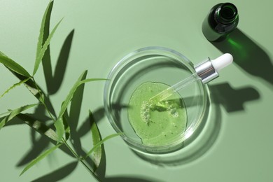 Photo of Petri dish with cosmetic product, bottle, pipette and leaves on green background, flat lay