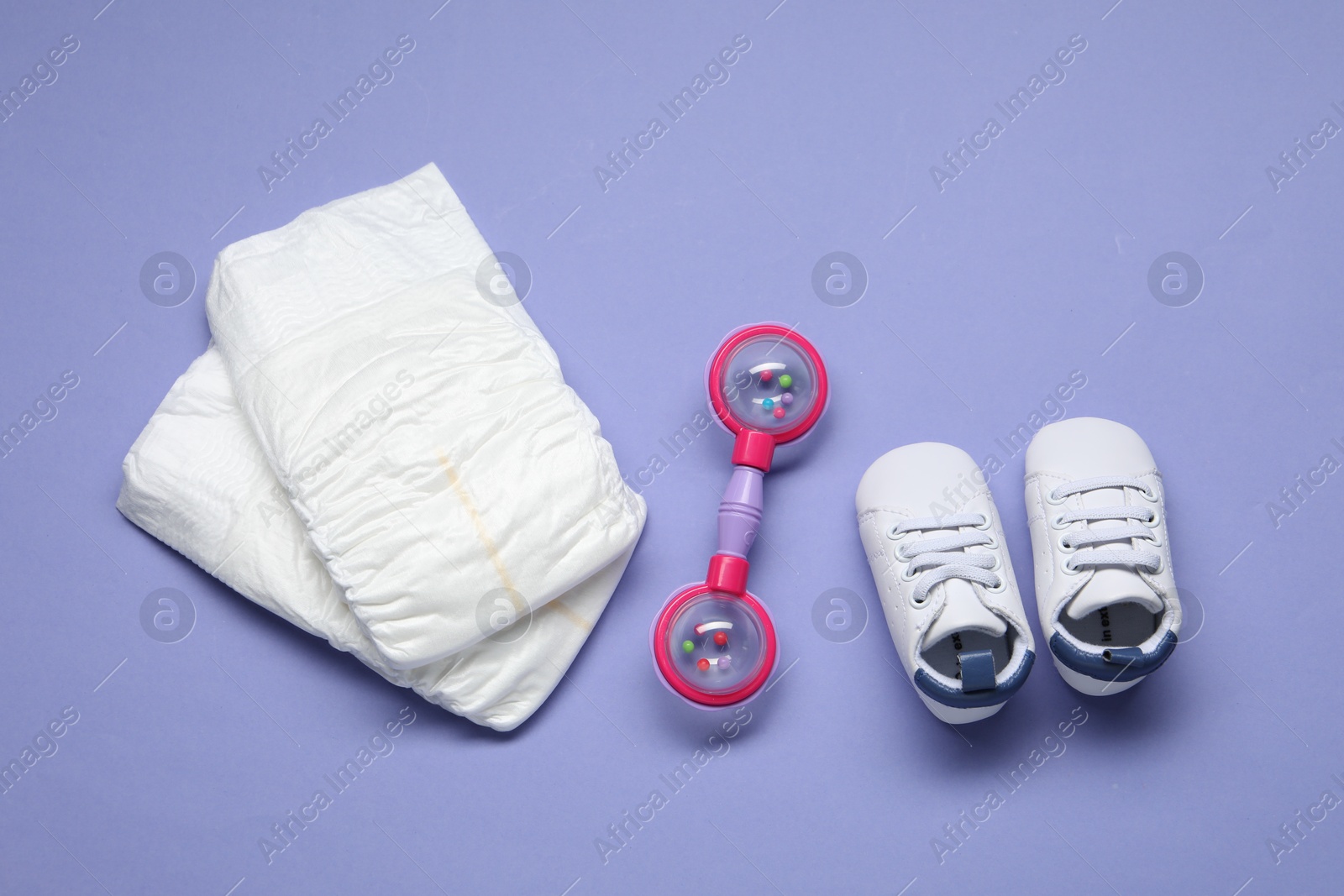 Photo of Colorful rattle, diapers and baby shoes on violet background, flat lay