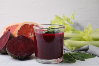 Photo of Tasty beetroot smoothie with basil in glass, fresh vegetables and grapefruits on light tiled table, closeup. Vegan drink