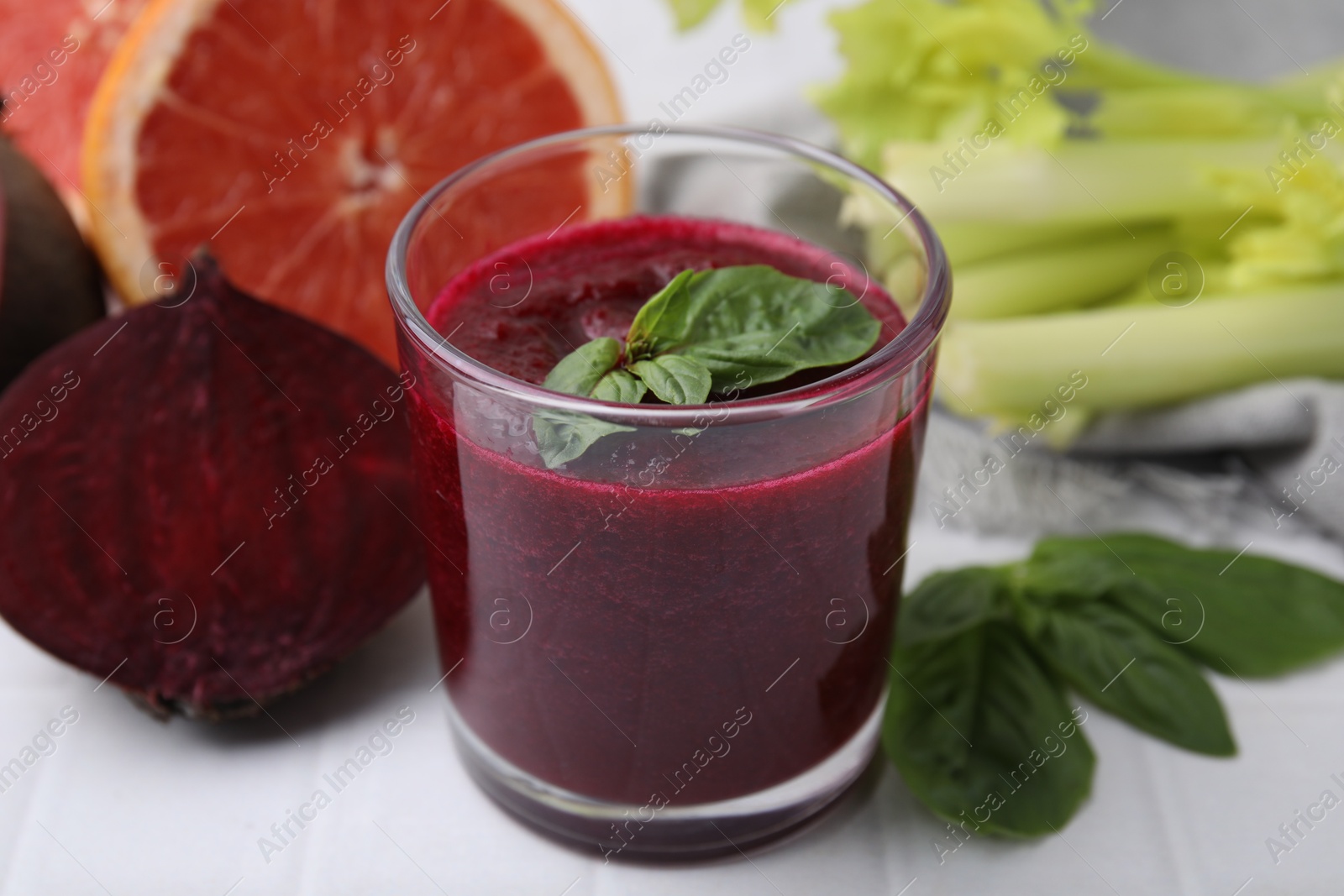 Photo of Tasty beetroot smoothie with basil in glass, fresh vegetables and grapefruit on light tiled table, closeup. Vegan drink