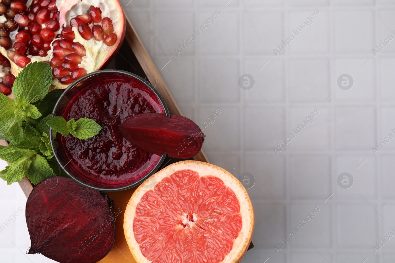 Photo of Glass of tasty beetroot smoothie, fresh vegetables, fruits and mint in wooden box on white tiled table, top view with space for text. Vegan drink