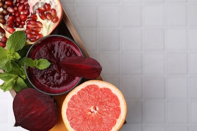 Photo of Glass of tasty beetroot smoothie, fresh vegetables, fruits and mint in wooden box on white tiled table, top view with space for text. Vegan drink