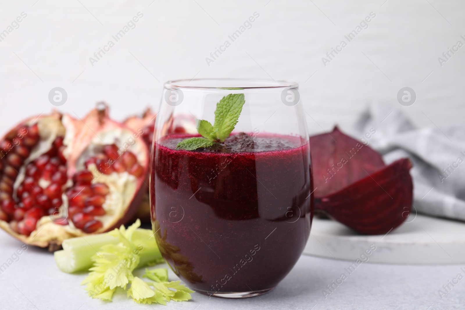 Photo of Tasty beetroot smoothie with mint in glass, fresh vegetables and pomegranate on light table, closeup. Vegan drink