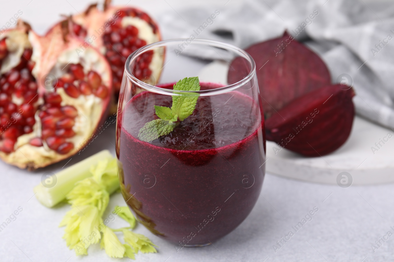 Photo of Tasty beetroot smoothie with mint in glass, fresh vegetables and pomegranate on light table, closeup. Vegan drink