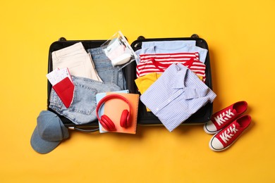 Open suitcase with traveler's belongings on yellow background, flat lay