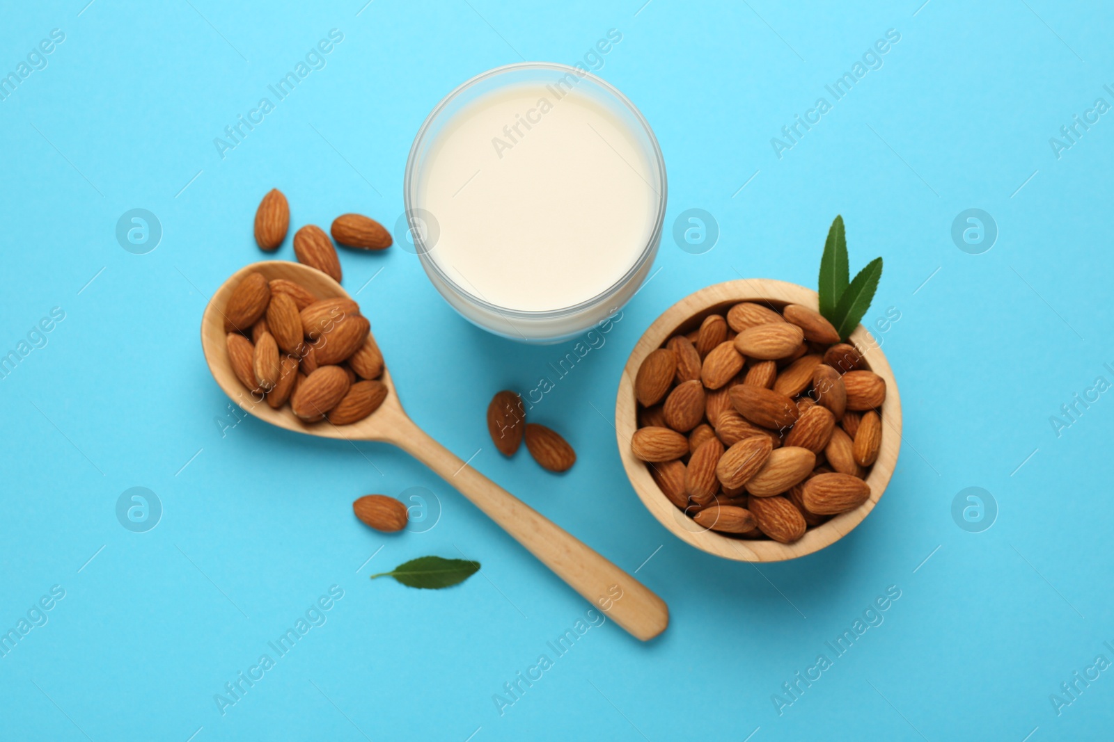 Photo of Fresh almond milk in glass, nuts, spoon and green leaves on light blue background, flat lay