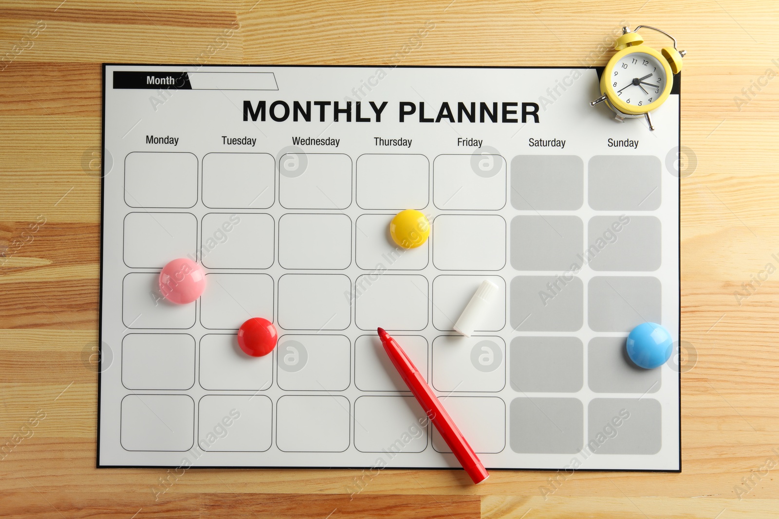 Photo of Timetable. Monthly planner, felt pen, drawing pins and alarm clock on wooden table, top view