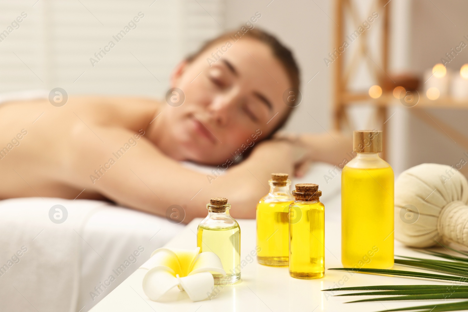 Photo of Aromatherapy. Woman relaxing on massage couch in spa salon, focus on bottles of essential oils, herbal bag and plumeria flower