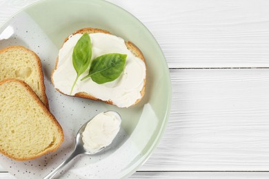 Pieces of bread with cream cheese and basil leaves on white wooden table, top view. Space for text