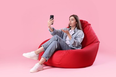 Emotional woman with smartphone having online meeting on red bean bag chair against pink background