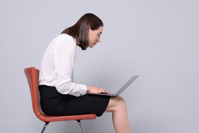 Woman with poor posture sitting on chair and using laptop against gray background, space for text