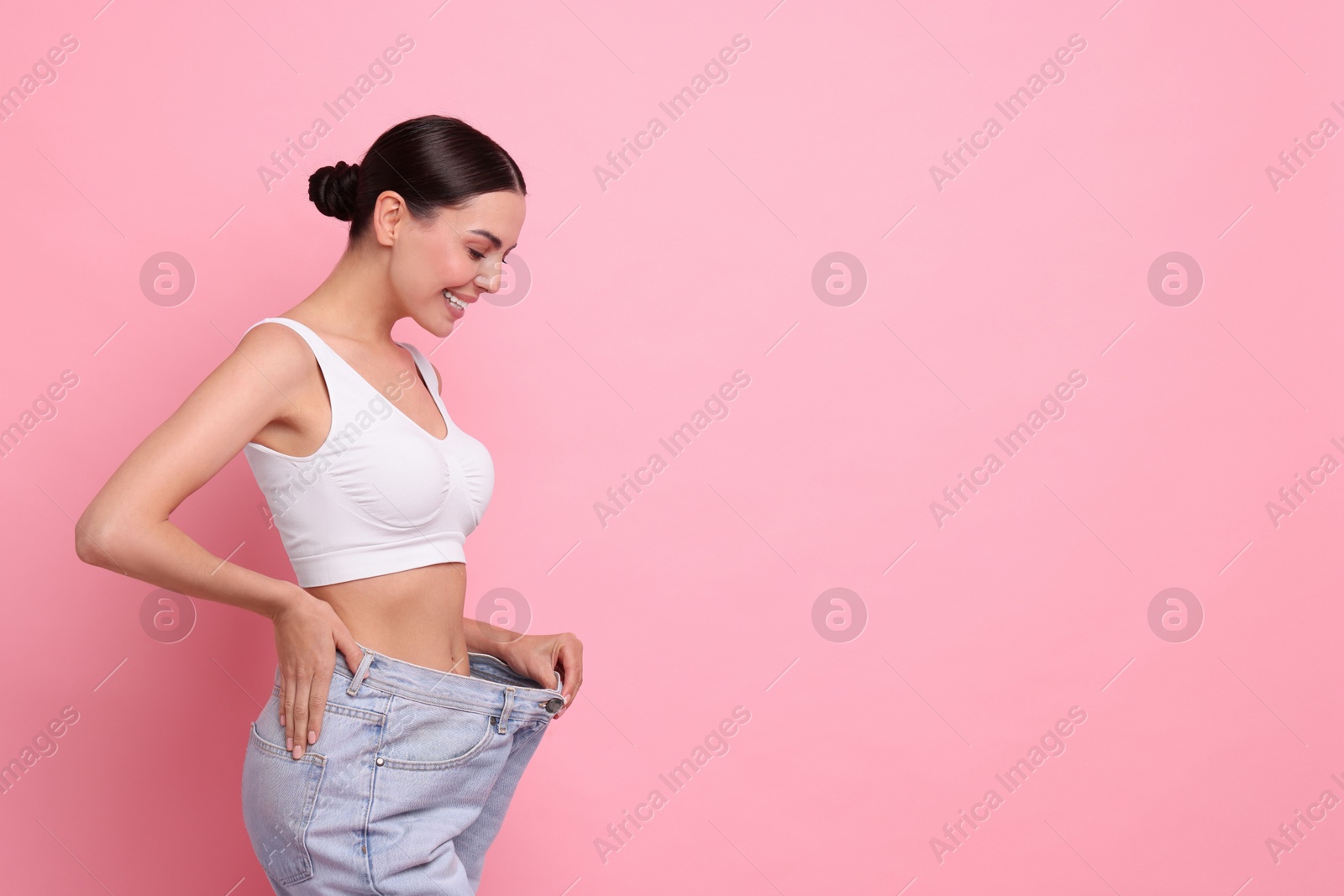 Photo of Diet and weight loss concept. Happy young woman in big jeans showing her slim body on pink background. Space for text