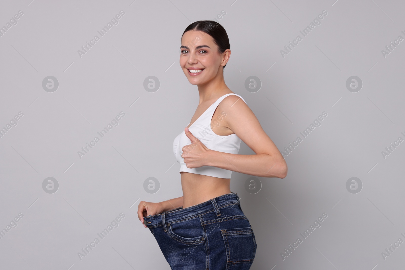 Photo of Diet and weight loss concept. Happy young woman in big jeans showing her slim body on light grey background