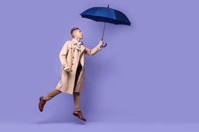 Photo of Young man with blue umbrella jumping on purple background, space for text