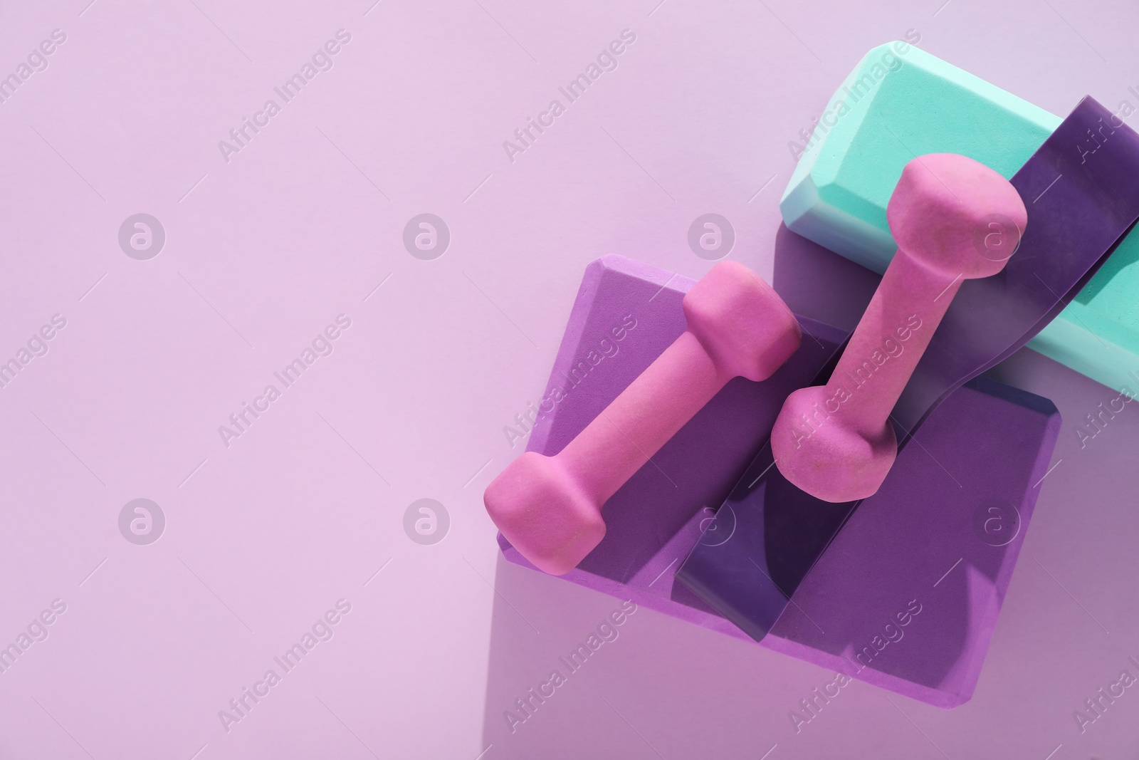 Photo of Two dumbbells, yoga blocks and fitness elastic band on violet background, flat lay. Space for text