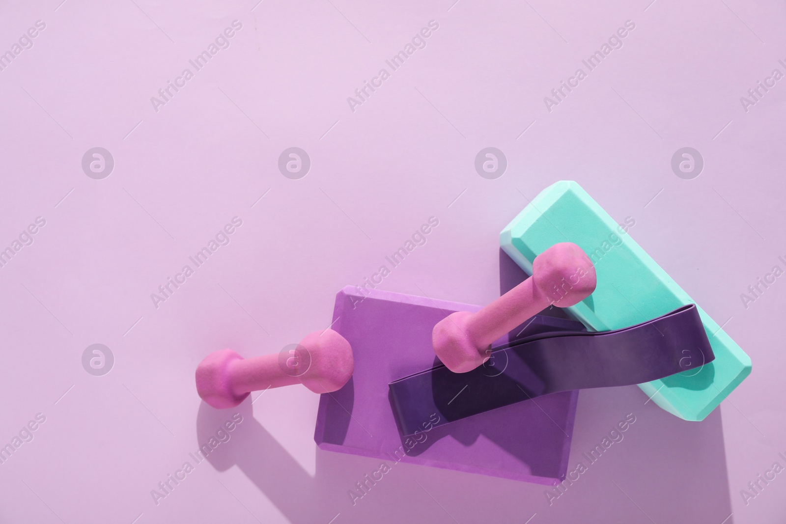 Photo of Two dumbbells, yoga blocks and fitness elastic band on violet background, flat lay. Space for text