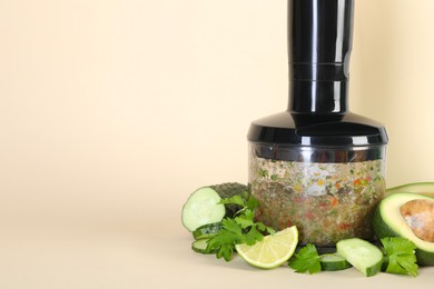 Photo of Hand blender with mixture of ingredients and fresh products on beige background, space for text