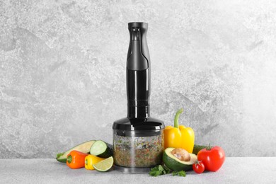 Hand blender with mixture of ingredients and fresh products on light grey table