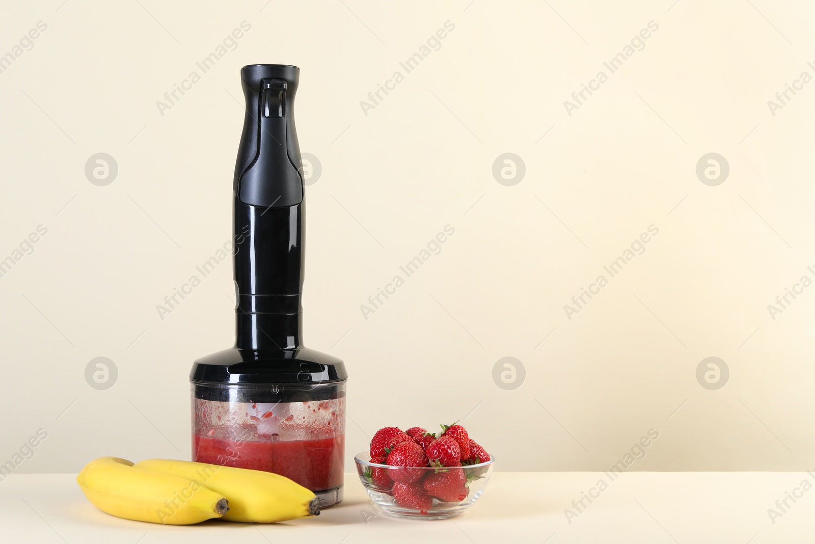 Photo of Hand blender with mixture of ingredients, banana and strawberries on beige background, space for text