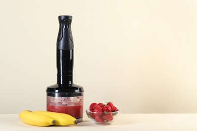 Photo of Hand blender with mixture of ingredients, banana and strawberries on beige background, space for text