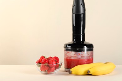 Hand blender with mixture of ingredients, banana and strawberries on beige background, space for text