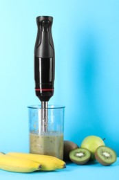 Photo of Hand blender with mixture of ingredients and fresh fruits on light blue background